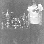 Black and white image of young woman athlete circa 1926 standing next to a table full of silver trophies she has won