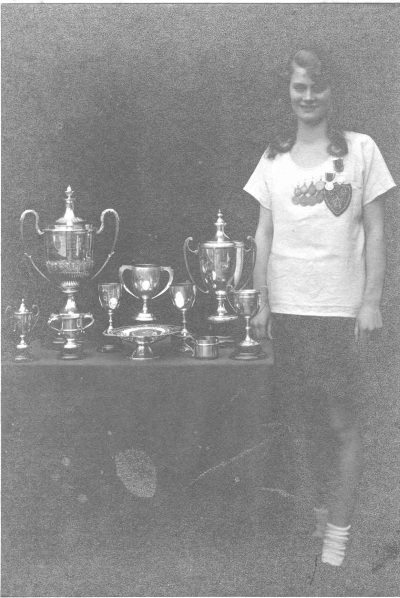 Black and white image of young woman athlete circa 1926 standing next to a table full of silver trophies she has won