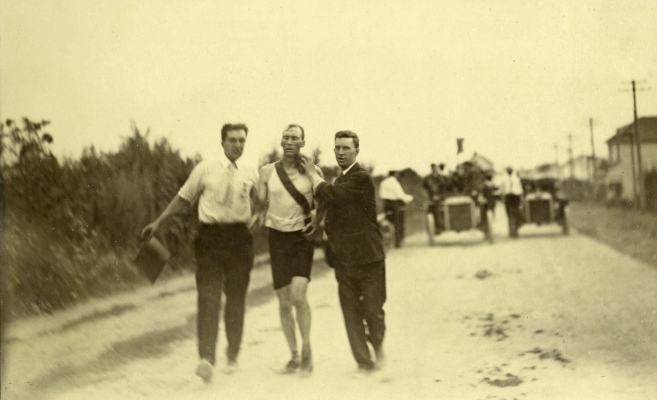 Black and white photograph from 1904 showing two men supporting a marathon runner between them on a dusty road, followed by two cars